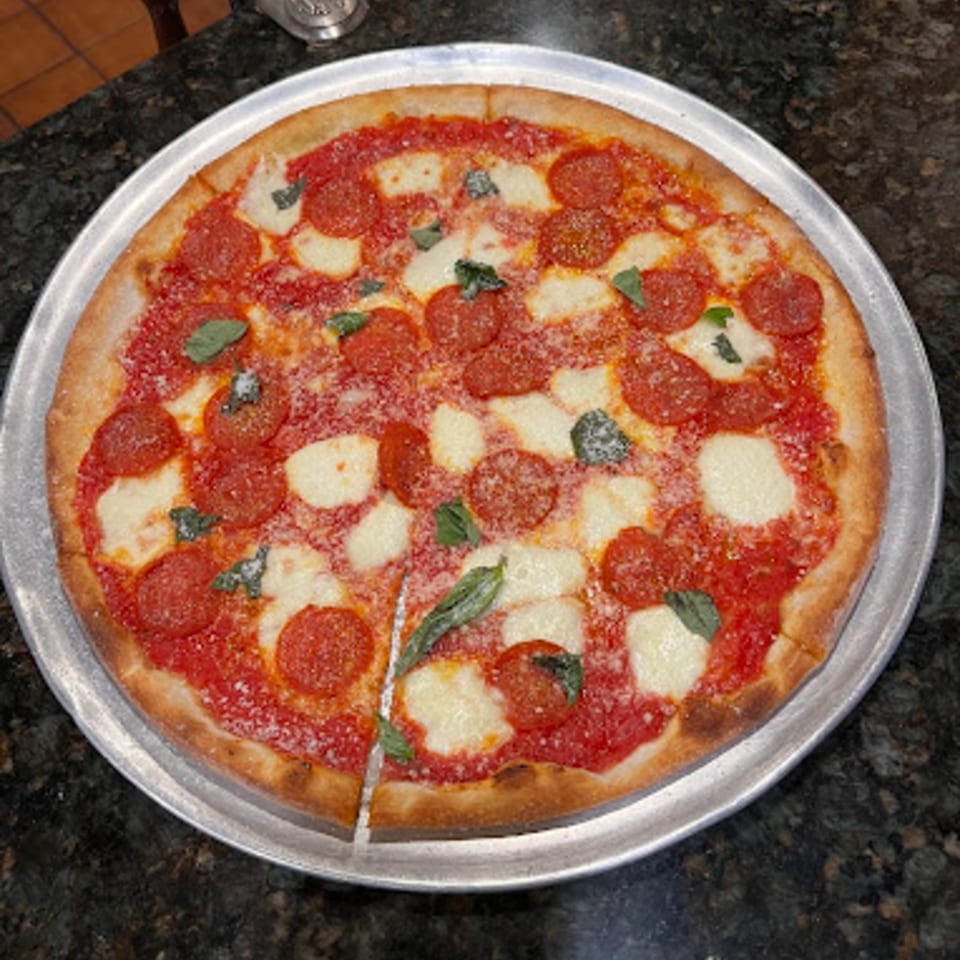 A whole pepperoni pizza with fresh mozzarella and basil on a metal tray, with one slice cut.