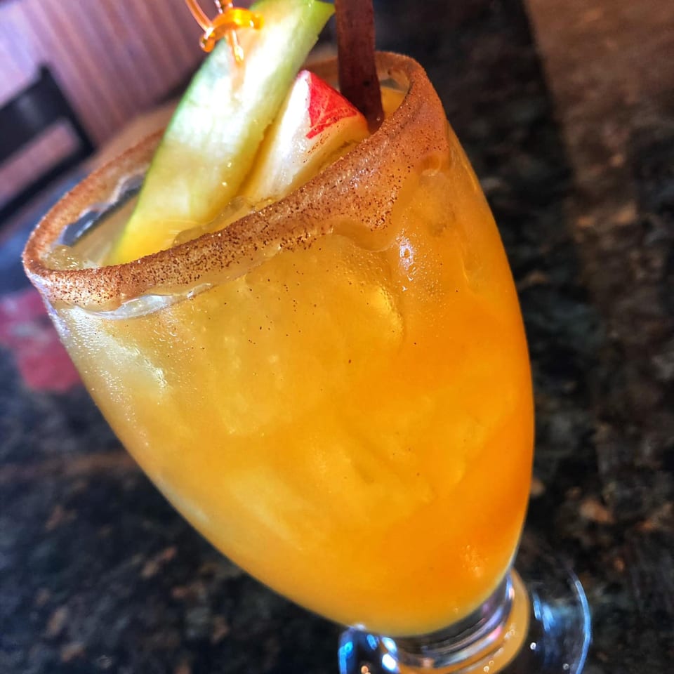 A vibrant orange cocktail in a sugar-rimmed glass, garnished with apple slices and a cinnamon stick.
